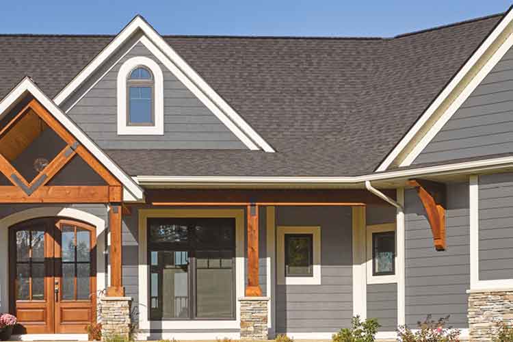 Picture of a house clad in gray siding, accented by natural wood columns in a craftsman style. The roof is a brownish-gray asphalt shingle. The home features several energy efficient cooling windows and doors all shining in the bright light of summer.
