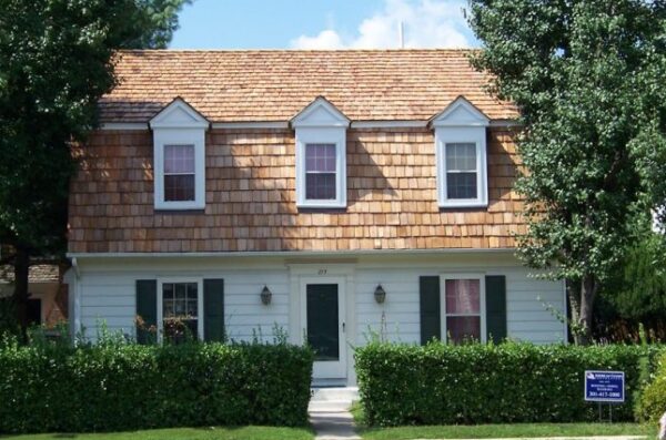 A well-maintained two-story home with cedar shake roofing and white siding, representing exterior renovations protected by warranties for defects in materials or workmanship. The photo was taken on a bright sunny day. The 2 large trees one on either side of the home are full of large dark green leaves. The box hedge across the front is green and healthy.