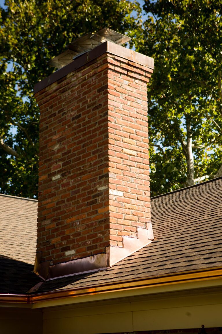 This image is custom copper chimney flashings on a brown colored asphalt roof system. The image depicts close up of a red brick chimney standing tall with the fresh custom copper flashing. photo was taken on a sunny day.