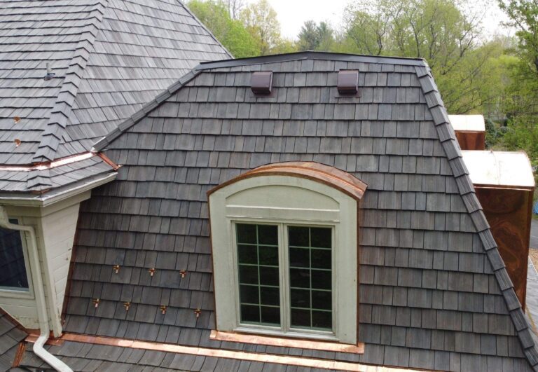 This image is of a Brava synthetic cedar shake system. The color is canyon gray. This is a close up of a portion of the roof which features a custom copper accent flashing around the new window. This photo was taken on a bright day with bright green trees behind.