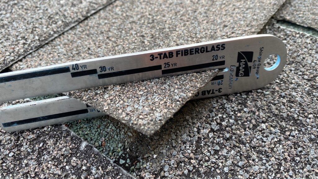 This image shows a close-up of a metal asphalt shingle gauge measuring a piece of gray asphalt shingle on a bright day. At American Custom Contractors this is just 1 of the vital tools we come with when we come out to perform a roof inspection. The tool shows the warranty of the shingle.