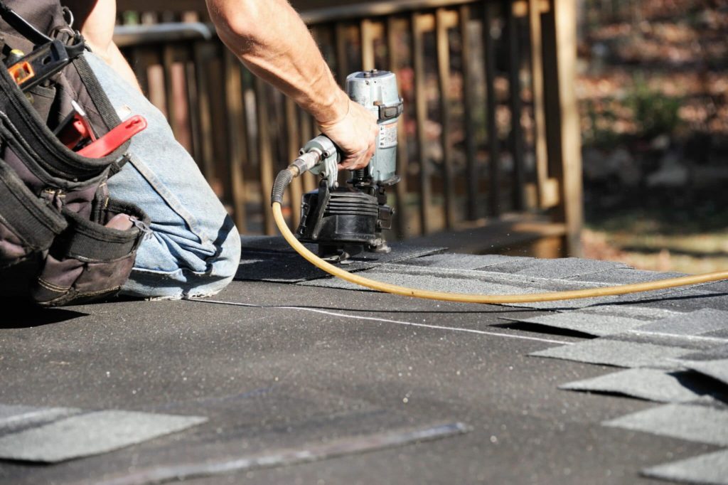 A roofer on his knees using the pneumatic nail gun to shoot nails in black asphalt roofing shing.