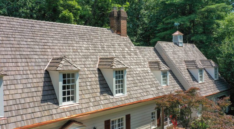 Photo of a Brava composite cedar shake roof tile system. Americna Custom Contractors installed this roof in 2023 and won an award for their stellar installation. The homeowner was so please with the outcome. The photo features the front elevation in a slight profile angle. It shows the custom copper gutter system and the natural color cedar shake tiles of the roof. 3 dormers are pictured.