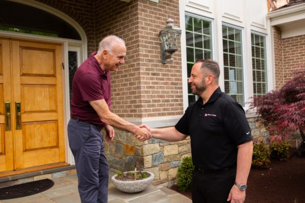 A business owner wearing a company uniform is greeted by a homeowner on his porch with a handshake.