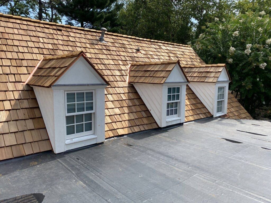 This photo showes a installtion in progress of a natural cedar shake roof. The image is taken from the roof of the 3 small dormer windows and the roof around complete. Photo taken on a sunny day with tall lush green trees in the background.