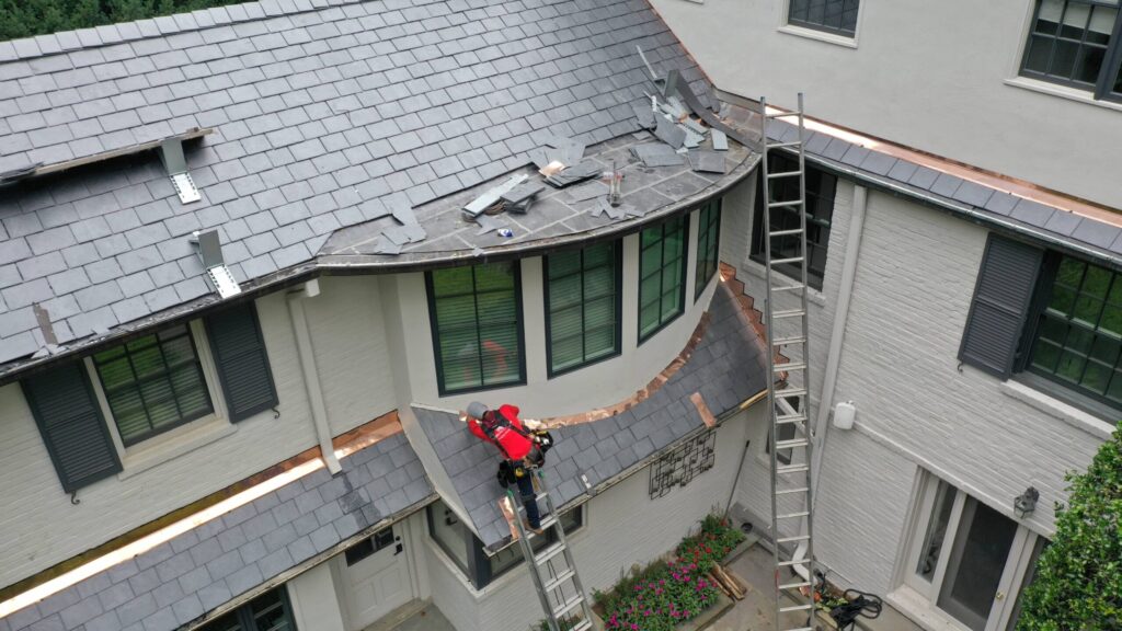 Roof installer on a ladder wearing a red shirt. They are installing natural slate tiles by hand, one by one on a large home in Maryland. We use this image to show a snapshot of the roof replacement process on a natural slate roof.