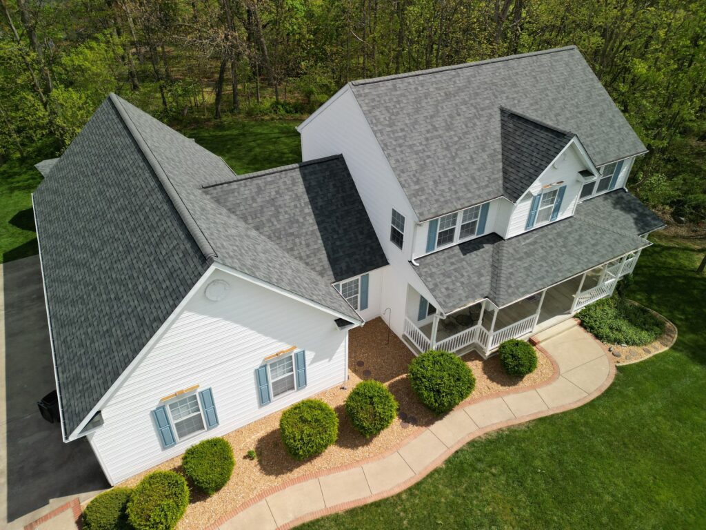 This drone image taken on a bright sunny day is of a farmhouse style home set in a pastoral setting. The home just recieved a roof replacement with a new Owens Corning Asphalt roof system in Estate Gray installed by American Custom Contractors. The home is surrounded by a well manicured yard with round green box hedge.