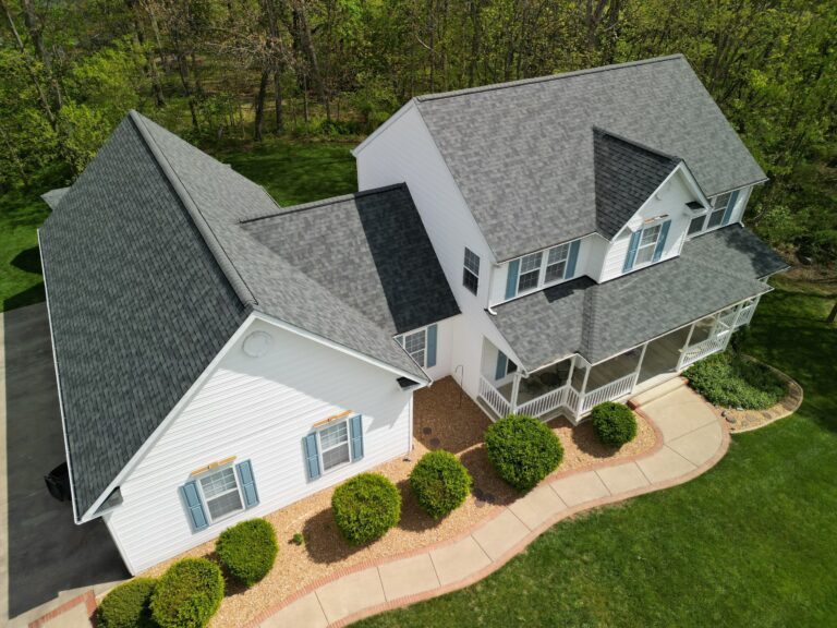 This drone image taken on a bright sunny day is of a farmhouse style home set in a pastoral setting. The home just recieved a new Owens Corning Asphalt roof system in Estate Gray installed by American Custom Contractors. The home is surrounded by a well manicured yard with round green box hedge.