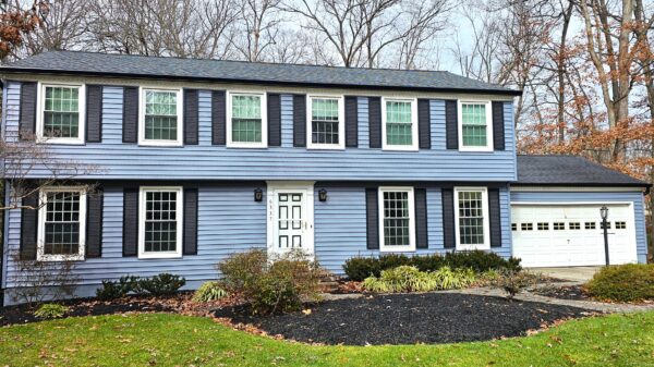 2-story home, with 6 windows on the 2nd level and 2 widows and a white door on the lower. The home is sporting brand-new blue vinyl siding and black shutters. The roof asphalt shingle is a dark onyx. The photo is taken on a sunny winter day. The grass is a bright green with no leaves on the trees behind the house.