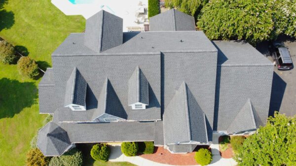 Drone shot of a large roof in Huntingtown Maryland. The roof features a gray shingle roof. The home has 7 dormer windows and a cupola. The photo was taken on a bright sunny day. The grass and trees are very green.