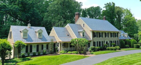 Large multiple-building home located in Potomac, Maryland. The home features natural black slate tiles on the roof, gleaming copper gutters and downspouts, and cream-colored vinyl siding with red shutters. The phot was taken on a bright sunny day and the grass and trees are a vivid green.