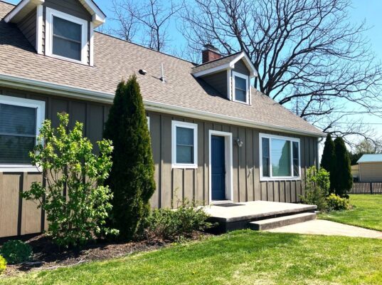 Photo taken at an angle of the front elevation. The single-story home features wood siding set in a vertical direction with 5 windows 2 are dormers set on a gray/brown asphalt shingle roof. The picture was taken on a bright sunny day and the grass is bright green.