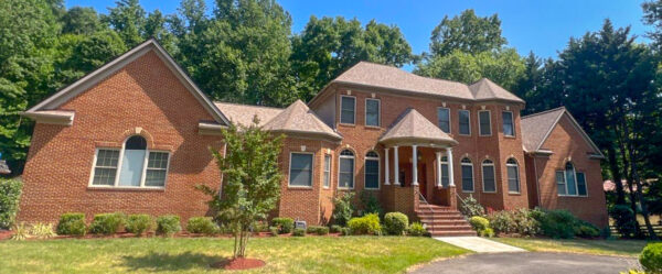 Front elevation of a large red brick 2-story sprawling home. the brick stairs lead up to a copula-style portico with 6 white columns. 20 large windows adorn the front of the home, along with a premium roof shingle in a varied brown shingle. Organized plantings run the full front of the building with a young green tree planted in the green grass.