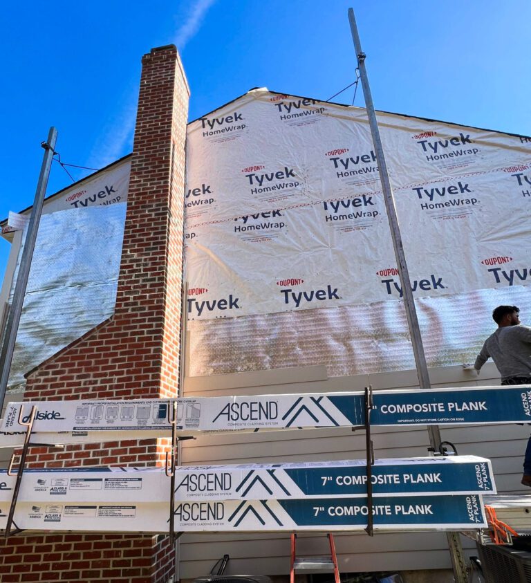 The photo shows the installation of Alside Ascend composite cladding over Low-E housewrap. This photo shows the side of the some with a tall red brick chimney and the scaffolding system that has beet up for the install. The low E is being installed over Tyveck house wrap. The boxes of Ascend siding are stacked up on the scaffolding ready for installation.