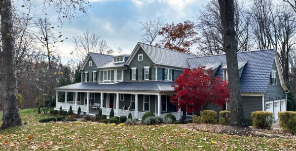 A large Potomac, Maryland home featuring James Hardie siding in a muted green color and a roof covered with EcoStar synthetic slate tiles. The house is two stories with a prominent front porch extending the length of the building. Surrounded by trees, including one with vibrant red autumn leaves, and a well-maintained garden, the home presents a charming, classic appearance.