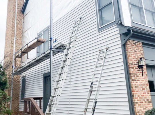 Picture of the side of a home in Falls Church Virginia, that is receiving an upgrade to its siding. The picture shows 2 ladders leaning up against the home and a tall scaffolding structure with a box of siding ready to install. The new white vinyl siding is installed halfway up.