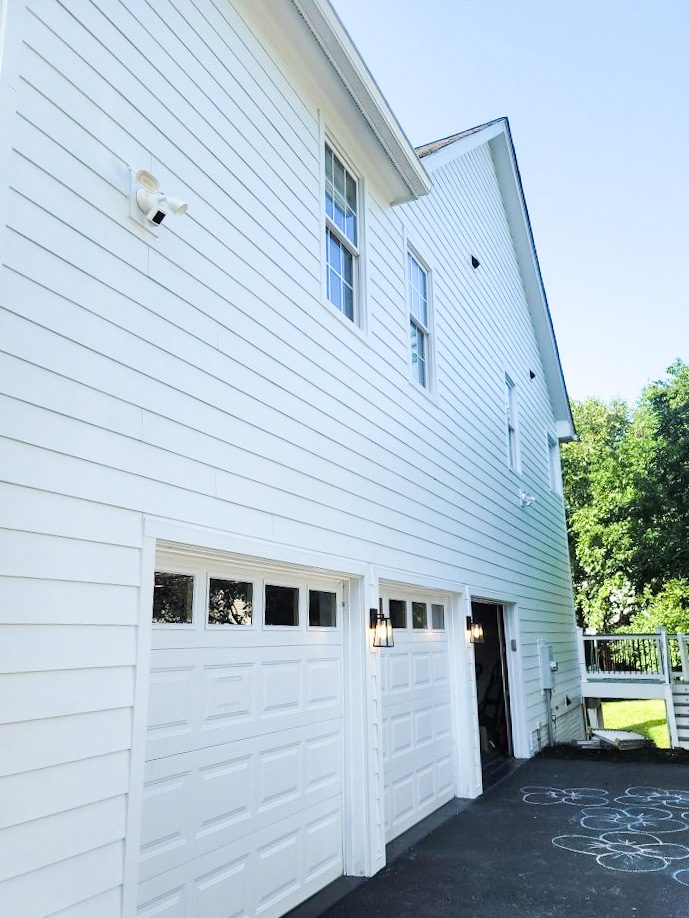 Picture of the side of a home in Gaithersburg, Maryland. This image features James Hardie plank fiber cement siding just installed in the color Alabaster white by Sherwin Williams. The contrast between the Black driveway and the bright siding looks fantastic. This photo was taken in the late afternoon and the result is a beautiful glowing sky and the dark green foliage of the trees contrasts the new light siding so nicely. And the addition of the black and glass carriage lights around the garage doors adds a touch of timeless elegance.