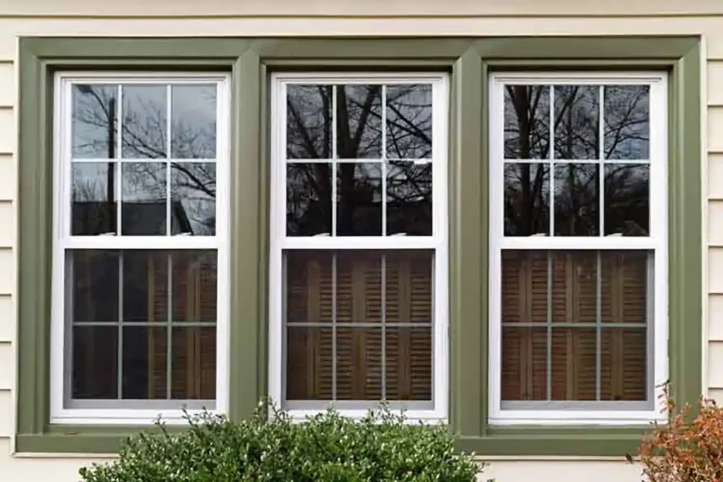 Picture of 3 windows on a home trimmed in a beautifull mossy green. The photo was teaken on a brigh day and you can see the reflection of bare tree branches in the glass. This image is used as a feature photo on the American Custom Contractors window & door replacement webpage.