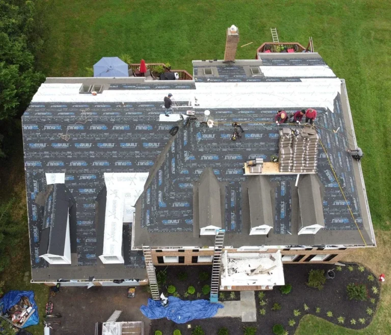 This image show American Custom Contractors installation crew on a roof that has had all its old roof material removed and a brand new underlayment installed as part of a repair and maintenance package. The image is a birds-eye-view on the entire roof. The install crew is wearing red shirts and safety harnesses.