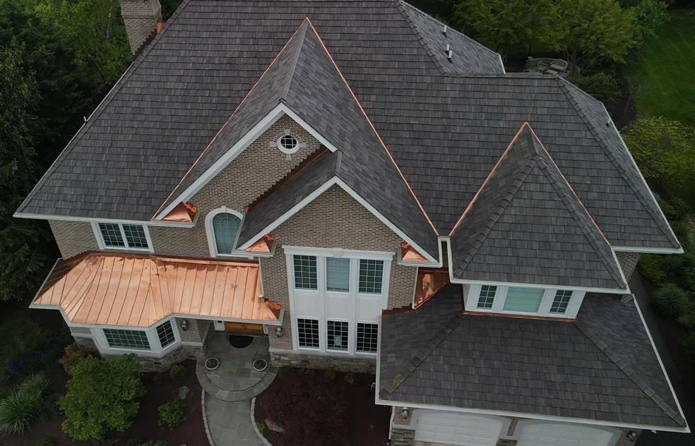 A bird's eye view of a large brick front home with a synthetic/composite cedar roof tile system installed. Accented by a standing seam copper accent roof and copper valleys. As well as replacement windows & doors. This home is in Potomac, Maryland