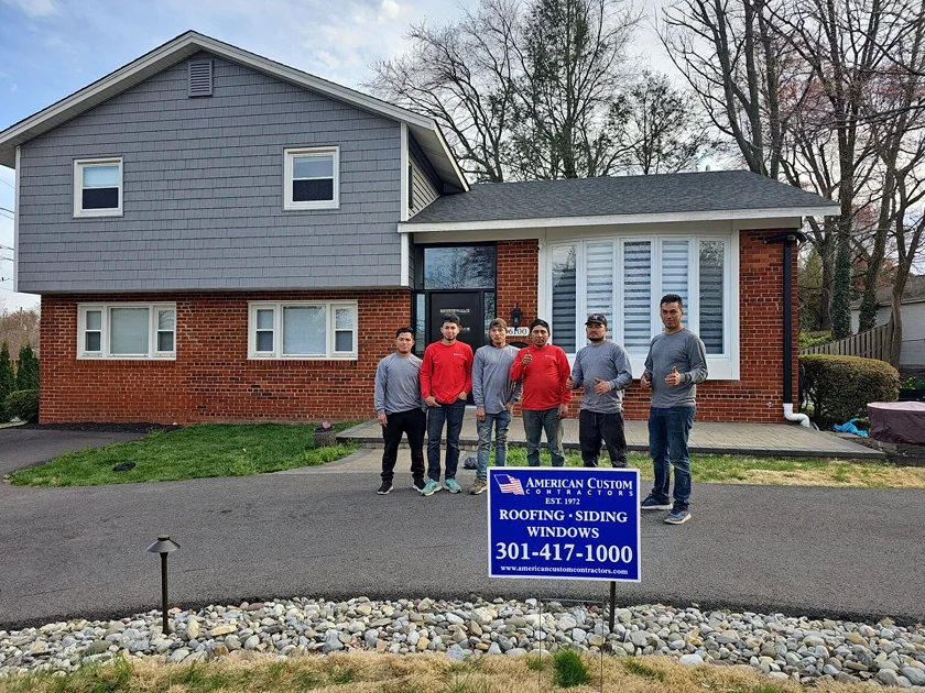 This Bethesda MD brick and gray vinyl siding ed home has a new GAF pewter gray asphalt. This picture shows the after shot of the home along with the installation crew dressed in red and gray American Custom Contractor shirts giving a thumbs up in front the the completed home.