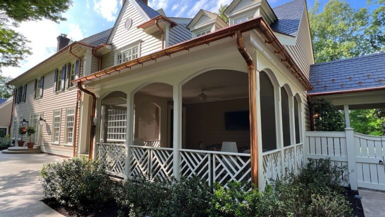 This photo is of the corner of a porch on a home in Potomac, Maryland taken on a bright day with blue sky in the background. The porch has a white lattice panels and white pillars. As well as custom copper gutters and downspouts installed by American Custom Contractors. There are green box hedges around the porch and cream colored paint on the porch celling.