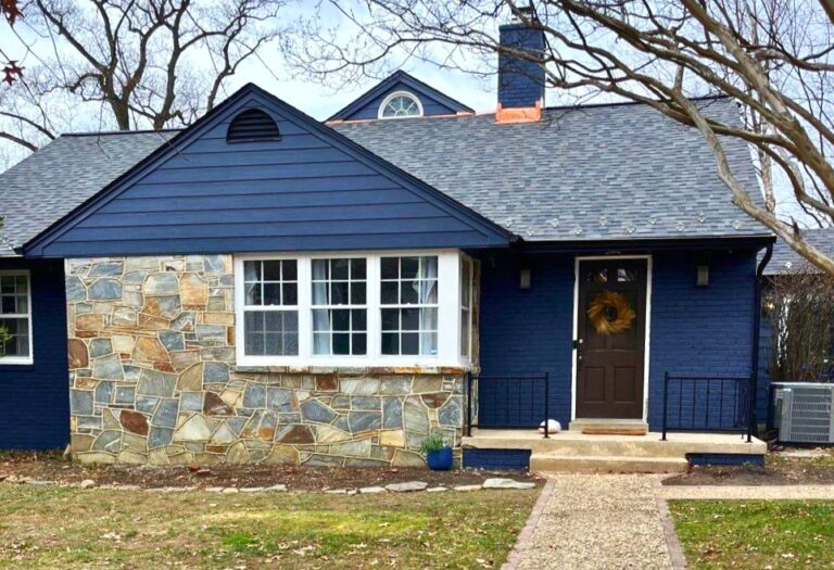 This image is of the front of a natural stone and siding single story home. American Custom Contractors just finished installing a Owens Corning Asphalt roof system and James Hardie cobalt blue siding. There bright copper flashings around the chimney. The photo was taken on bright sunny day. The trees in the background are bare of leaves.