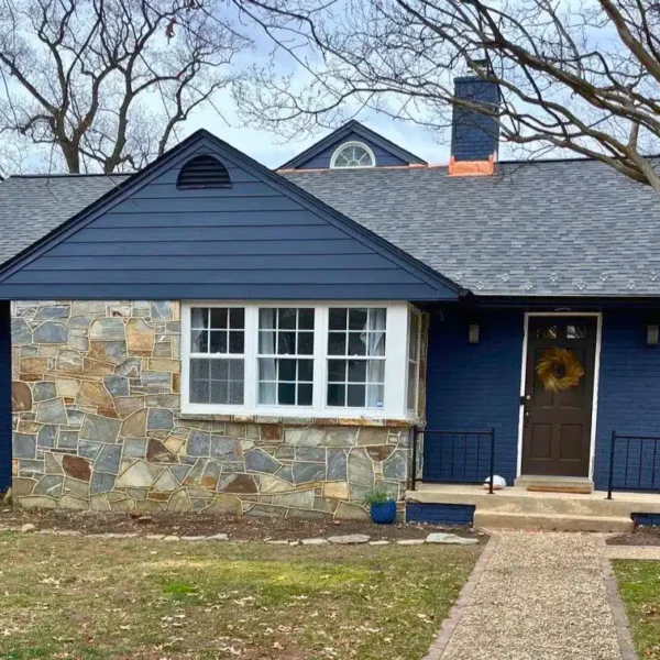 A beautifully renovated home in Arlington, VA, featuring James Hardie siding in a deep navy blue color, complemented by a natural stone accent wall. The house has a newly installed Owens Corning shingle roof in a dark gray tone, enhancing its modern yet classic appeal. The exterior also includes a navy blue brick facade, a brown front door with a wreath, and black metal railings. The renovation was completed by American Custom Contractors, showcasing high-quality craftsmanship and durable materials. The front yard features a stone walkway, potted plants, and a well-maintained lawn with bare trees in the background.