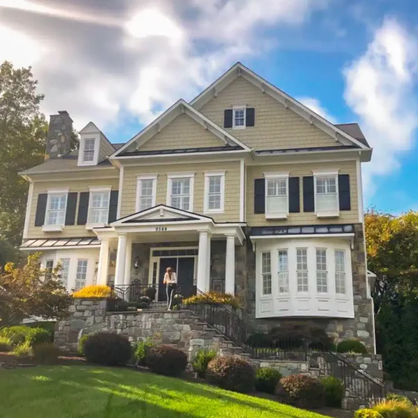 A beautiful custom-built home in Rockville, MD, featuring James Hardie siding in a beige tone with white trim and black shutters. The house has a stone foundation, a welcoming front porch with white columns, and a well-manicured landscape. The renovation was completed by American Custom Contractors, showcasing expert craftsmanship and attention to detail. A person is standing at the entrance, adding a sense of scale to the grand exterior.