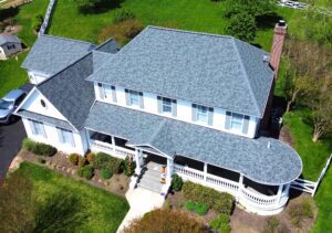 This image features a drone shot of a farmhouse style home with a large white porch that ends with a round coupula style roof on the right corner of the front of the home. It also features a newly installed Owens Corning asphalt roofing system in the color estate gray recently installed by American Custom Contractor after a large hailstorm that hit the area.