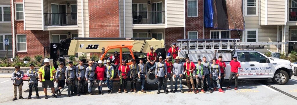 This a an image of a large group of construction workers standing together in front of a construction lift. They are wearing various safety harness equipment and are ready to get to work installing a new flat roof and siding on the apartment building shown in the background.