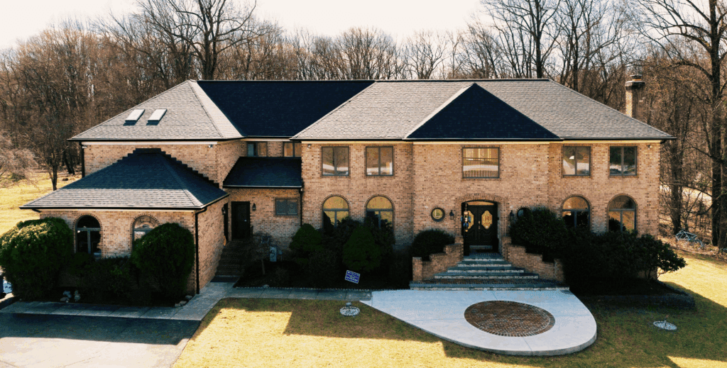 This photo of a large 2 story sandy colored brick home taken by drone represents work done by American Custom Contractors in Ellicotte Cirt MD. The home features a new Owens Corning dark colored roof with the name Peppercorn. The large trees areound are bare of any leaves and the grass is a faded winter brown/green. The sun is shining but the sky is a cloudless gray/blue.