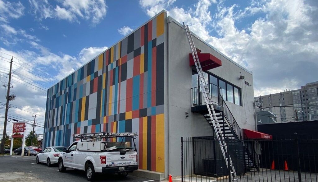 This photo is of a 2 story commercial building with a flat roof. There are ladders leaned up against the building and an American Custom Contractors truck with a tool box and ladder rack. They are replacing the flat roofing system.
